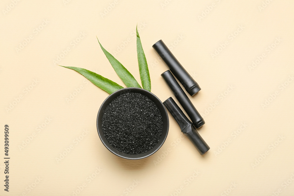 Bowl of activated carbon powder and black bamboo sticks on color background