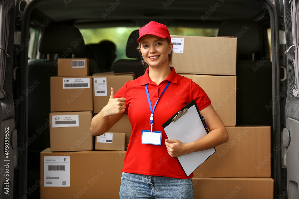 Female courier of delivery company showing thumb-up near automobile with parcels