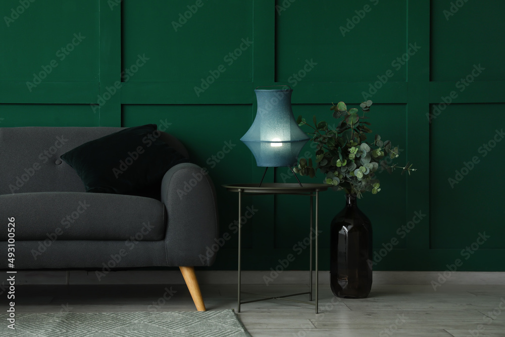 Glowing lamp on coffee table, vase with eucalyptus and sofa near green wall