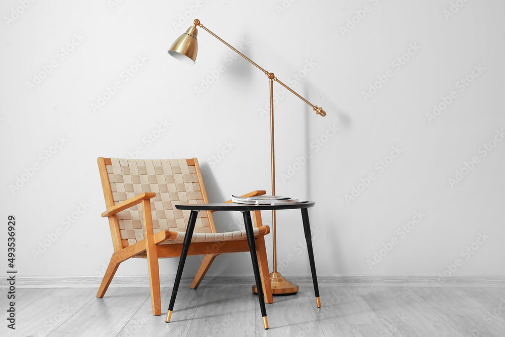 Comfortable armchair, table and floor lamp near light wall in room interior