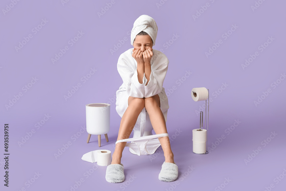 Young woman in bathrobe suffering from diarrhea while sitting on toilet bowl against color backgroun