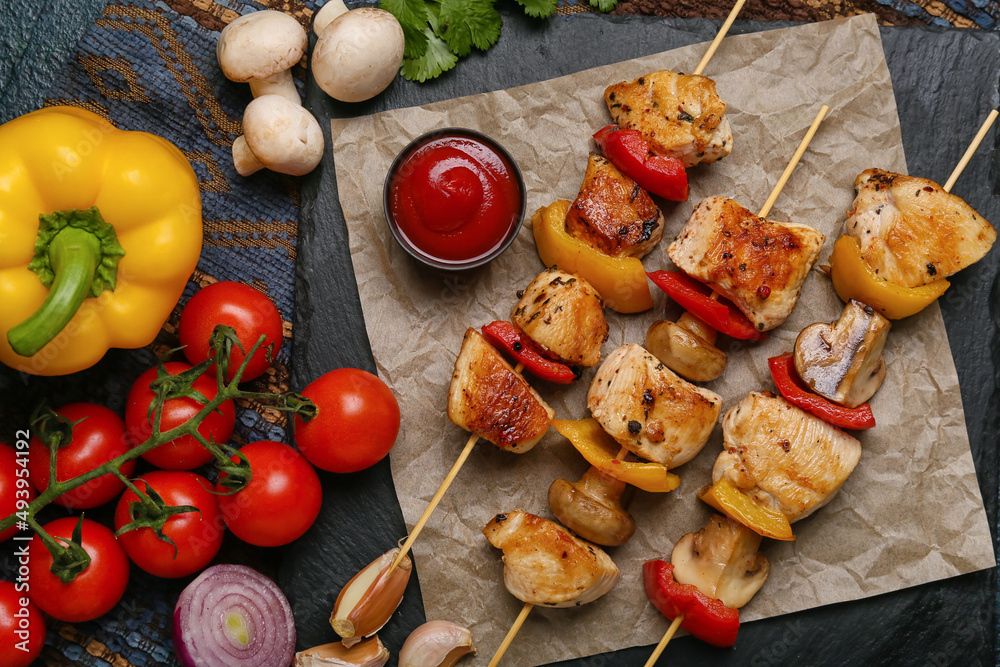 Board of grilled chicken skewers with vegetables and sauce on black background