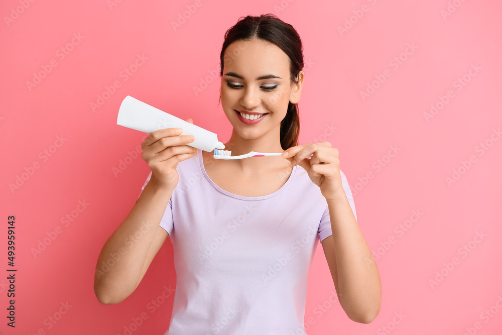 Young woman applying tooth paste onto brush on pink background