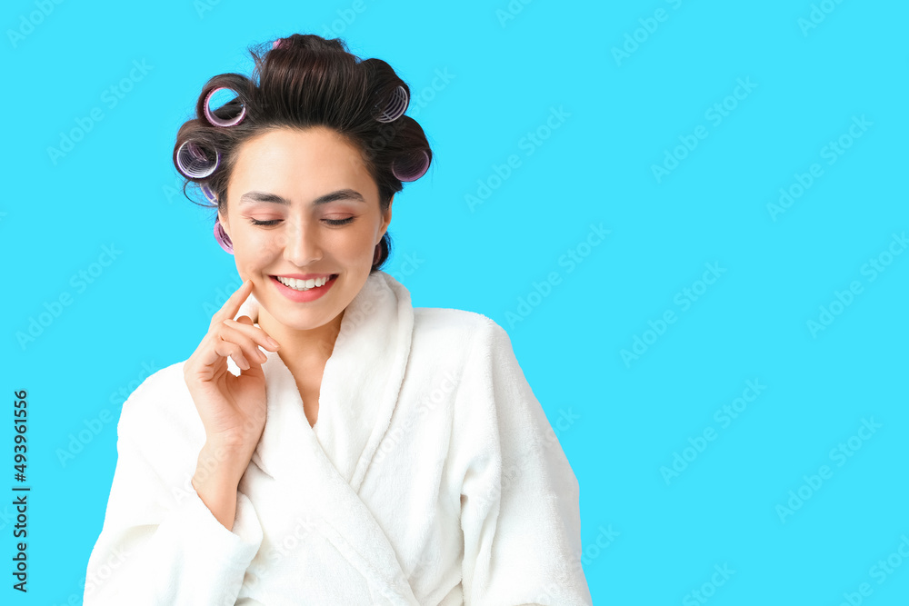 Beautiful young woman in bathrobe and hair curlers on blue background