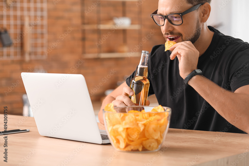 Handsome man with laptop drinking beer and eating chips in kitchen