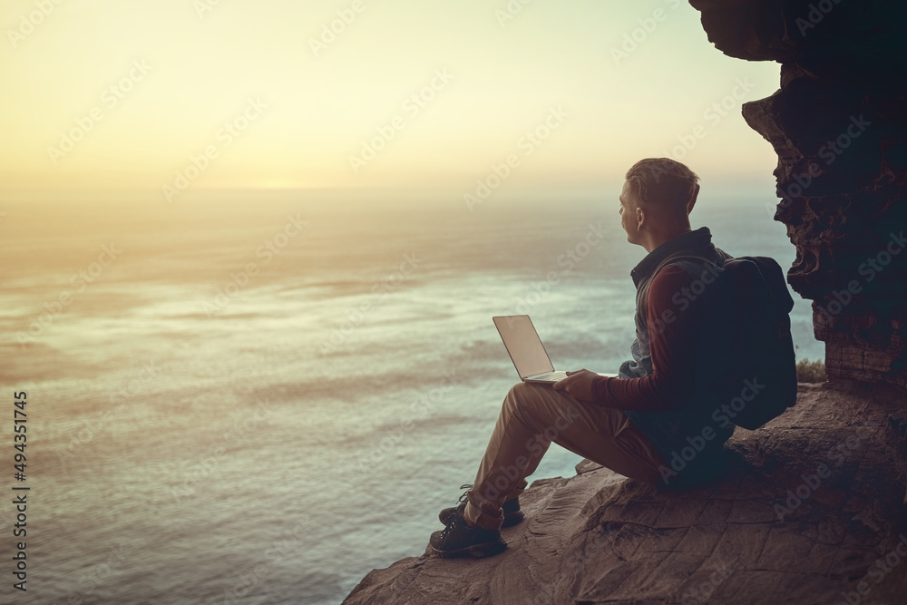 Connecting to the world. Full length shot of a young man using his laptop while sitting on a cliff i