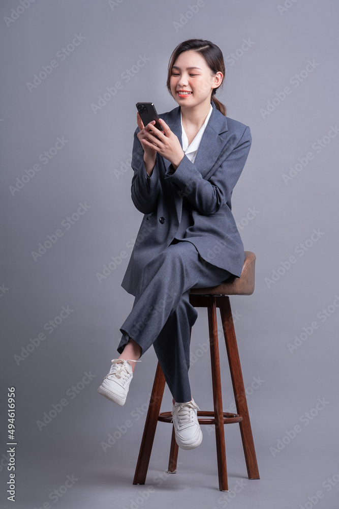 Young Asian businesswoman sitting on chair and using smartphone on gray background