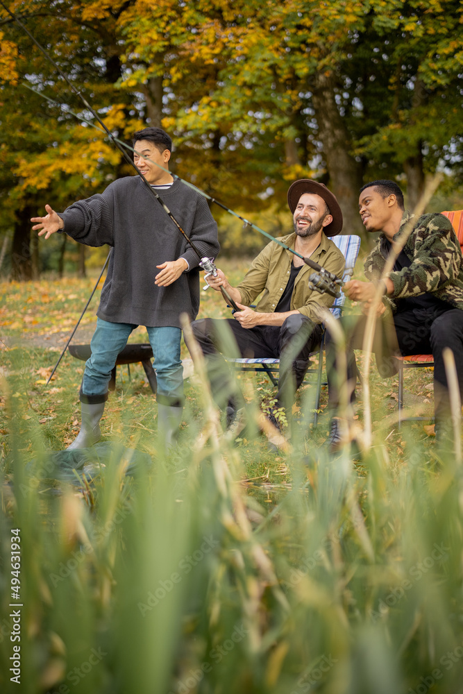 Pleased multiracial male friends resting and fishing on river or lake coast. Concept of leisure, hob