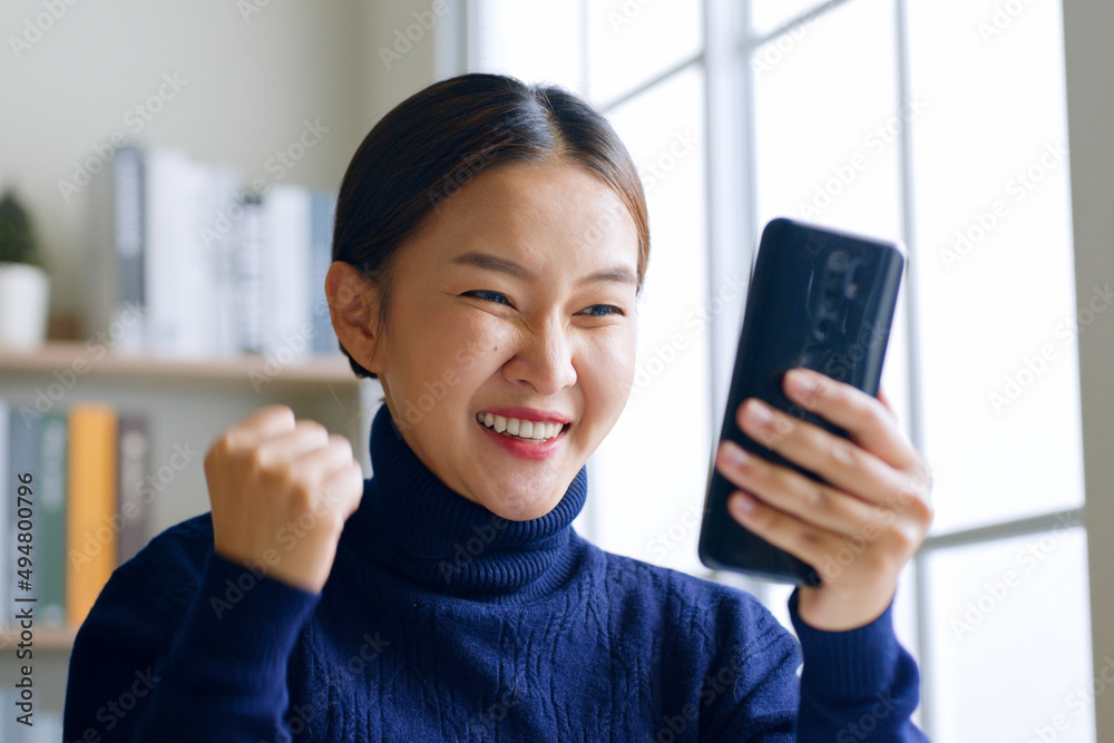 Young Asian woman using smartphone showing winner gesture, glad, excite at home