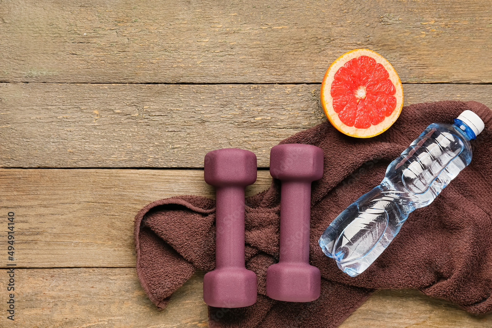 Stylish dumbbells, towel, bottle of water and grapefruit on wooden background