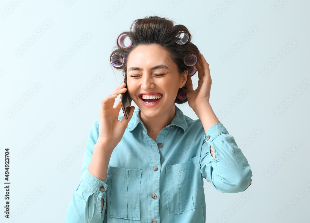 Laughing young woman in hair curlers talking by phone on light background