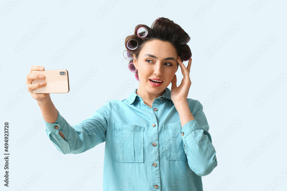 Beautiful young woman in hair rollers taking selfie on light background