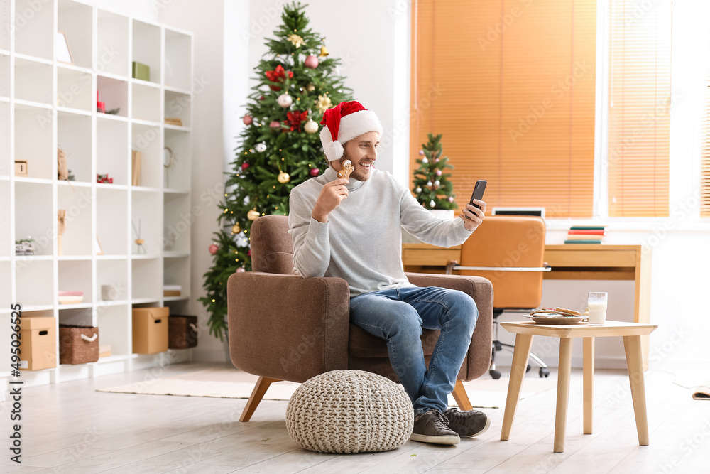 Young man in Santa hat with Christmas cookie taking selfie at home