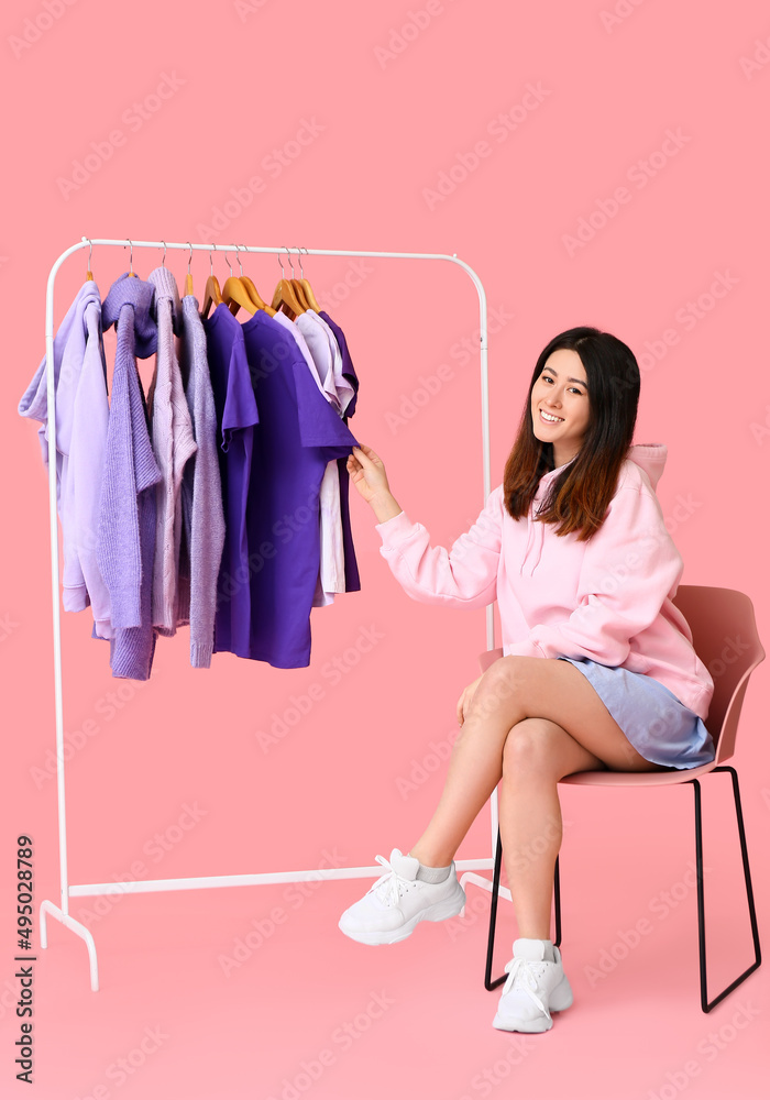 Young Asian woman sitting in chair and rack with clothes on pink background