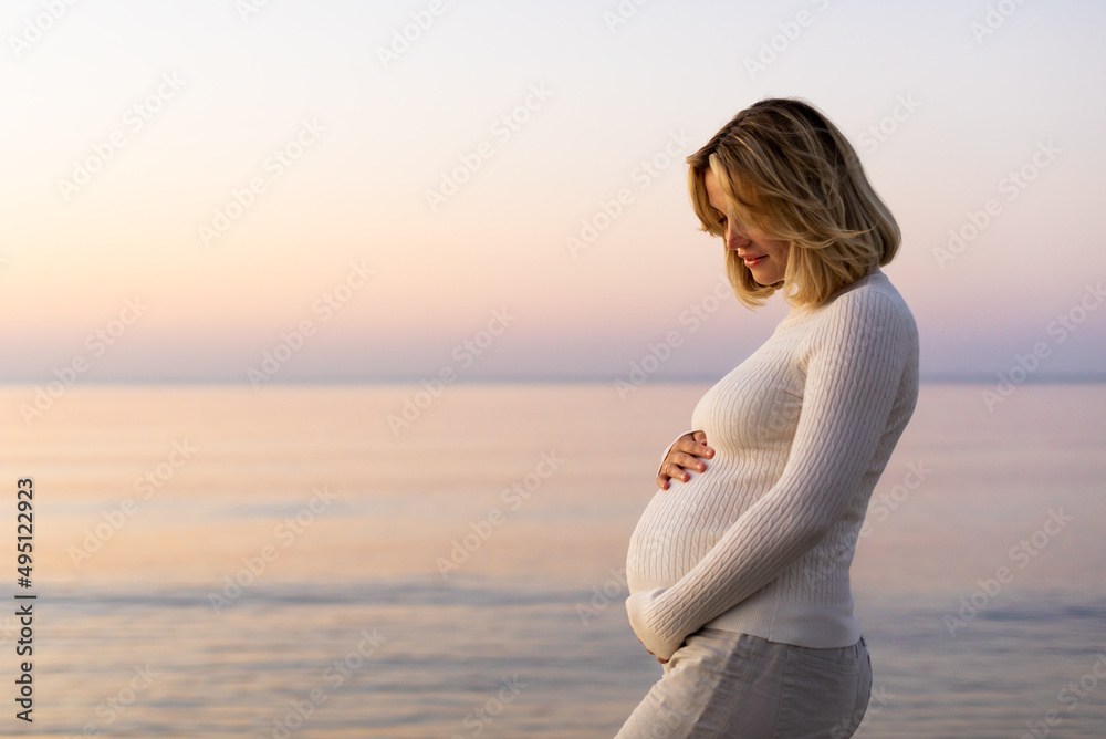 Charming pregnant woman walking on calm sea shore at evening