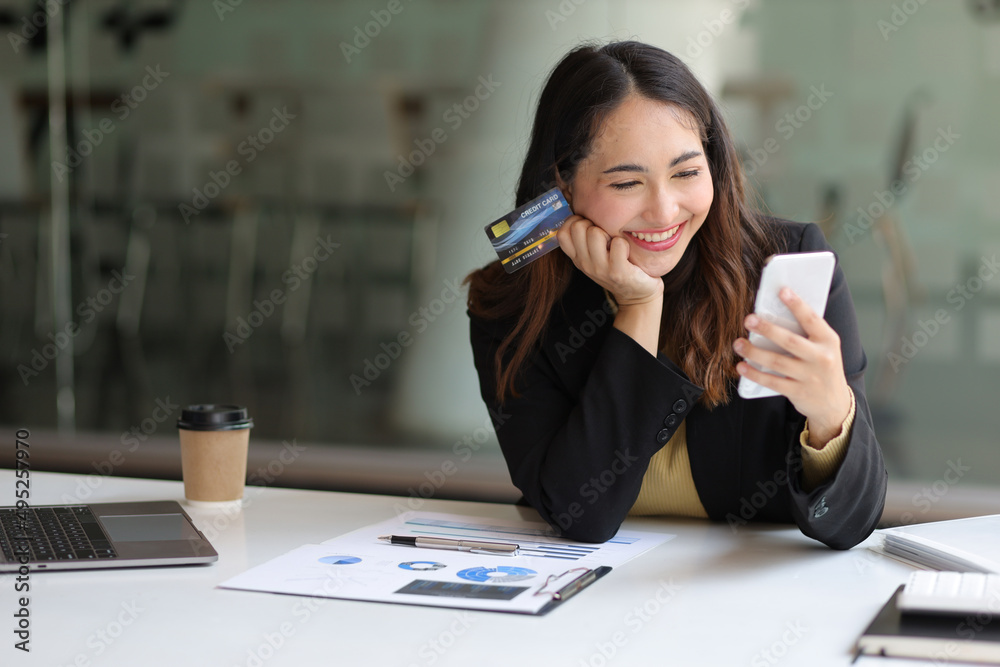 Cheerful businesswoman in office holding credit card and using mobile phone. Woman checking credit c