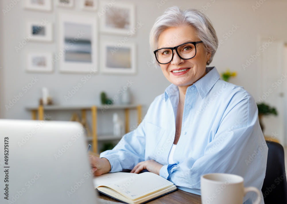 Mature happy businesswoman working at laptop, reading documents and take notes in home office