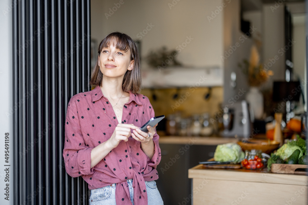 一个年轻快乐的女人站在厨房里用手机的肖像。带着pho的家庭主妇