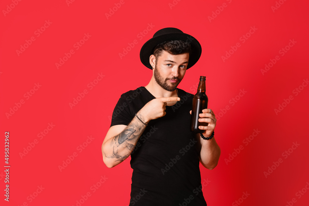 Stylish young man pointing at bottle of beer on red background