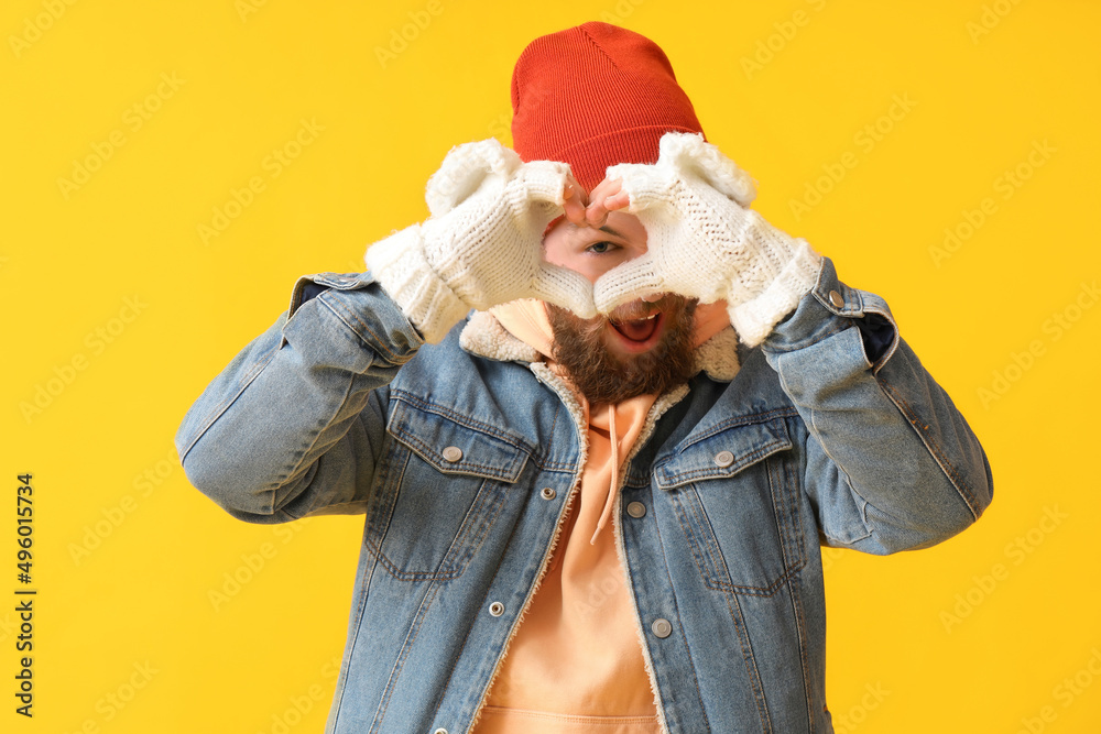 Young man making heart shape with his hands in warm gloves on yellow background