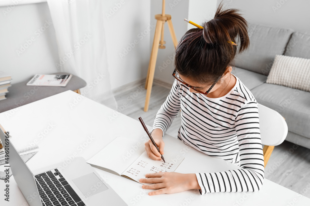 Little Asian girl writing in copybook during online lesson at home