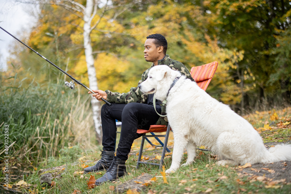 一名拉丁男子坐在椅子上，在河流或湖泊海岸的Maremmano Abruzzese Sheepdog附近钓鱼。Con