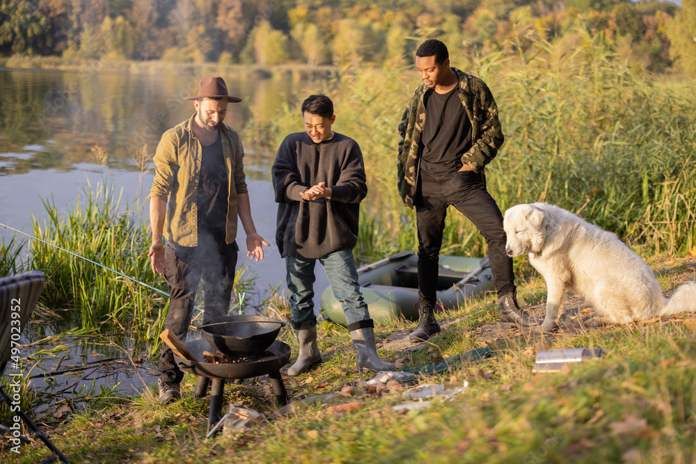 Multiracial group of male friends talking and cooking Ukha on fire. Men resting and fishing on lake 