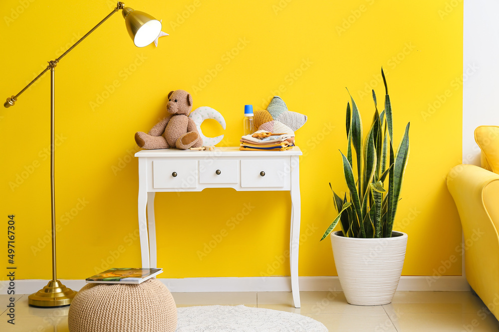 Table with baby clothes, toys, lamp and houseplant near yellow wall