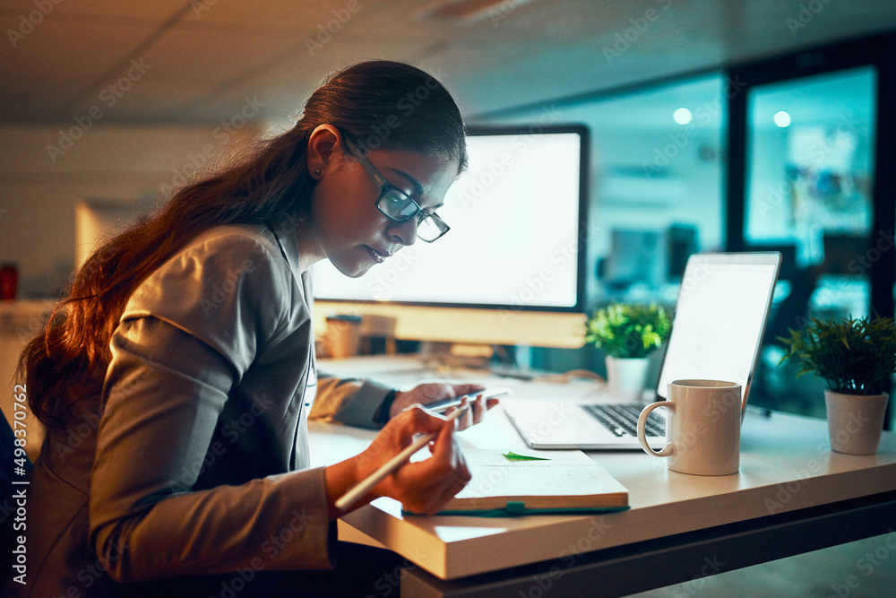 Writing down what she does as she does it. Shot of a young businesswoman working late in the office.