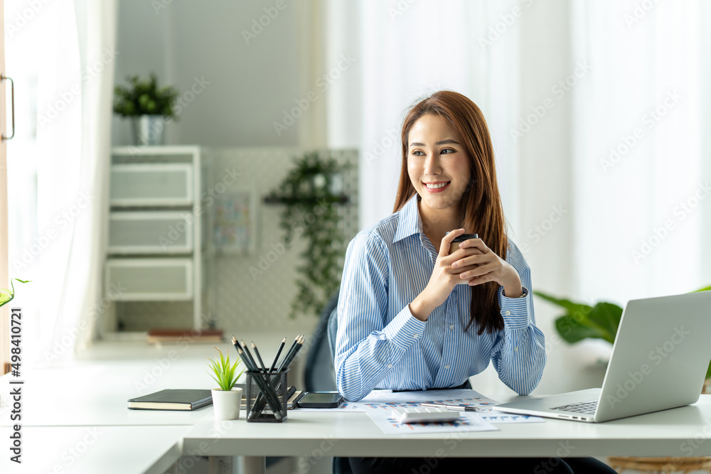 亚洲商务女孩在她的工作站上微笑着拿着咖啡杯。