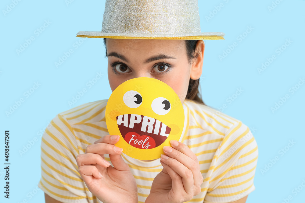 Young woman holding paper smile with text APRIL FOOLS on blue background