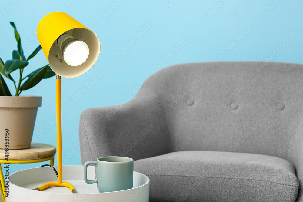 Table with stylish lamp and cup near armchair in interior of living room