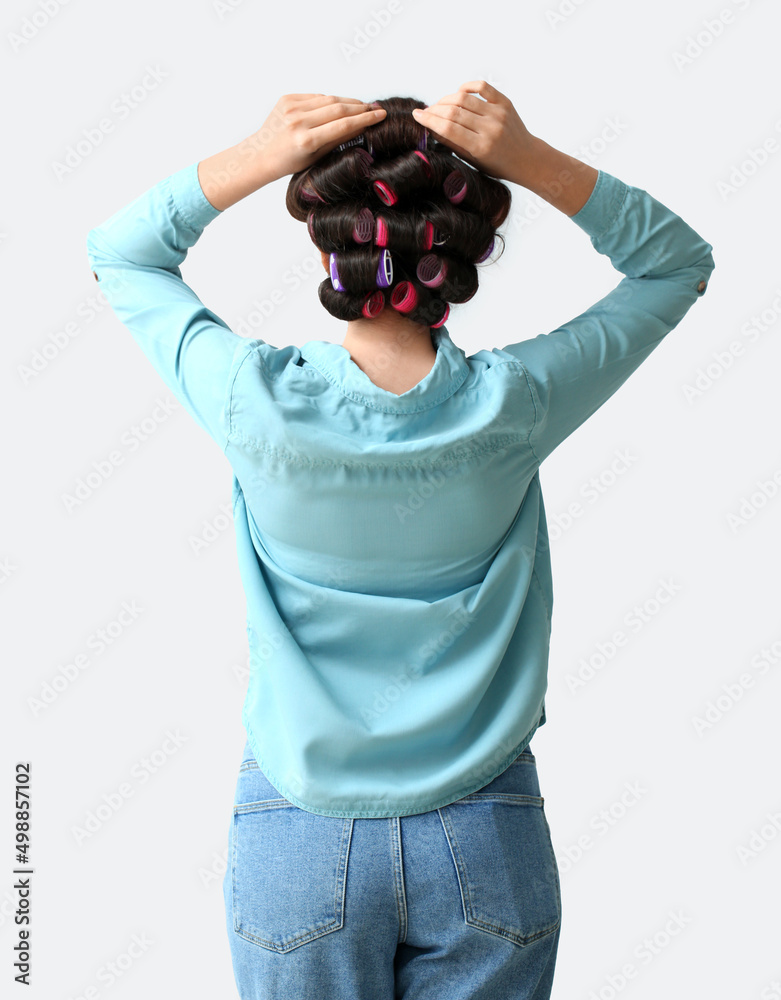 Beautiful young woman in hair rollers on light background, back view