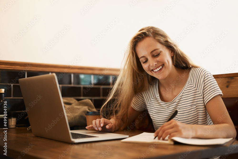 Getting ready to ace that test. Shot of a happy young student using her laptop to study at a cafe ta