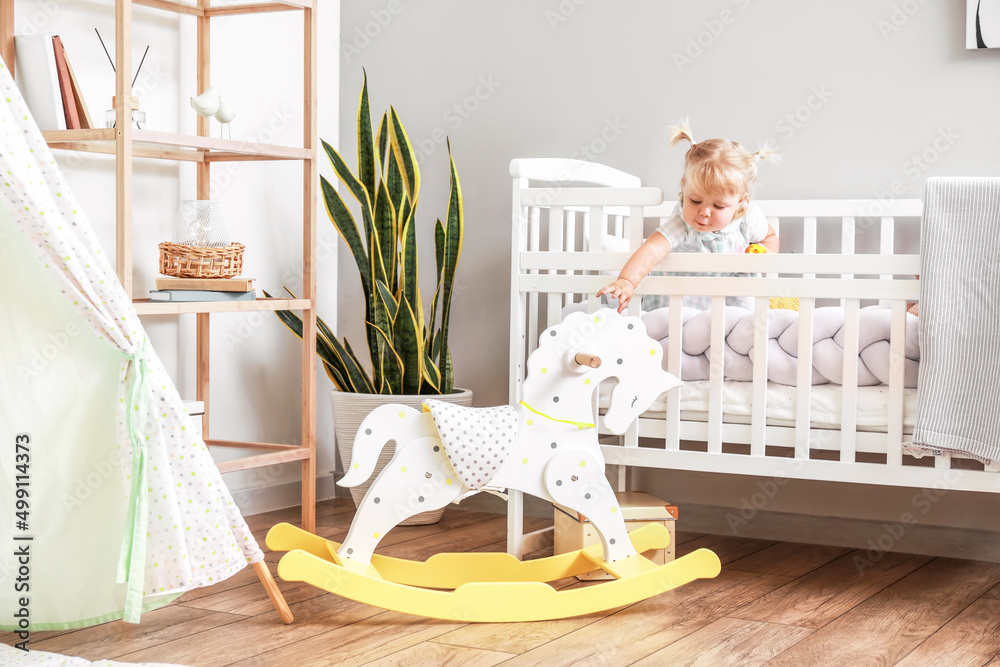 Adorable baby girl in bed near rocking horse in room