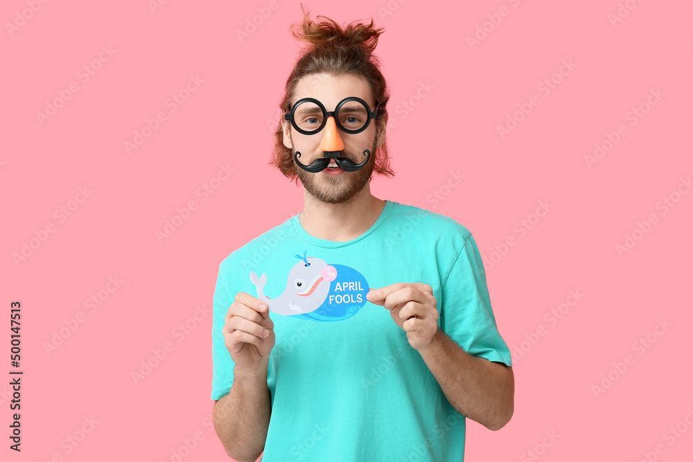 Funny young man in disguise and with card on pink background. April fools day celebration