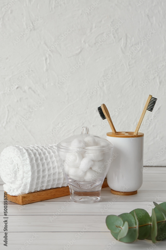 Bowl with cotton balls, clean towel and toothbrushes on light wooden table