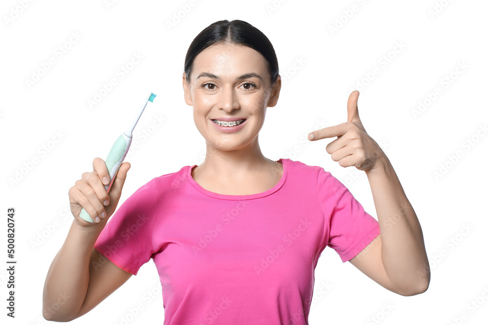 Beautiful woman with dental braces pointing at electric toothbrush on white background