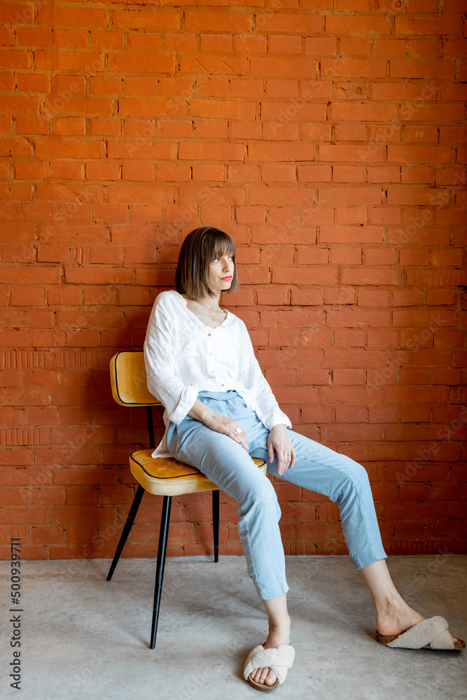 Full length portrait of a young woman dressed casually sits relaxed on chair on brick wall backgroun