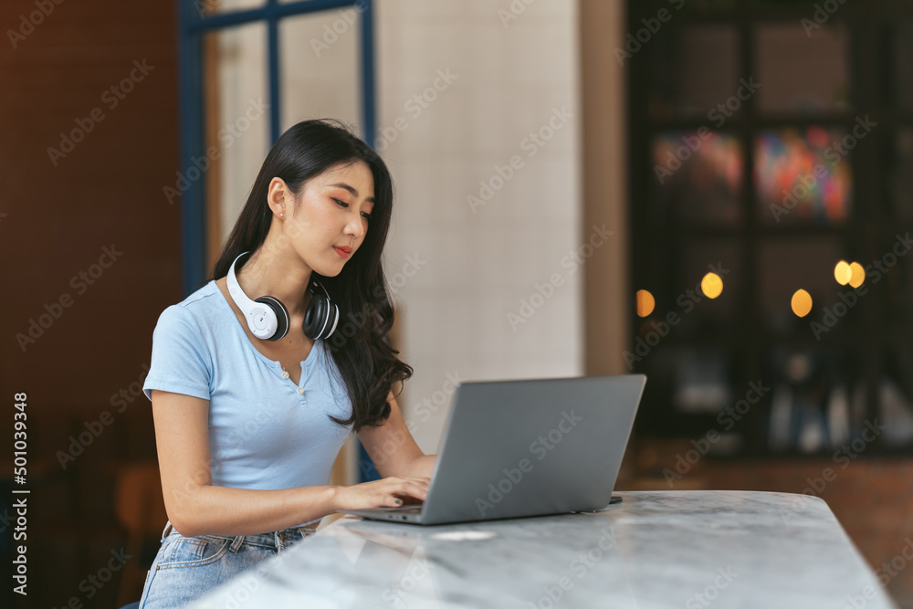 Asian young female student sitting at the table, using headphones when studying.