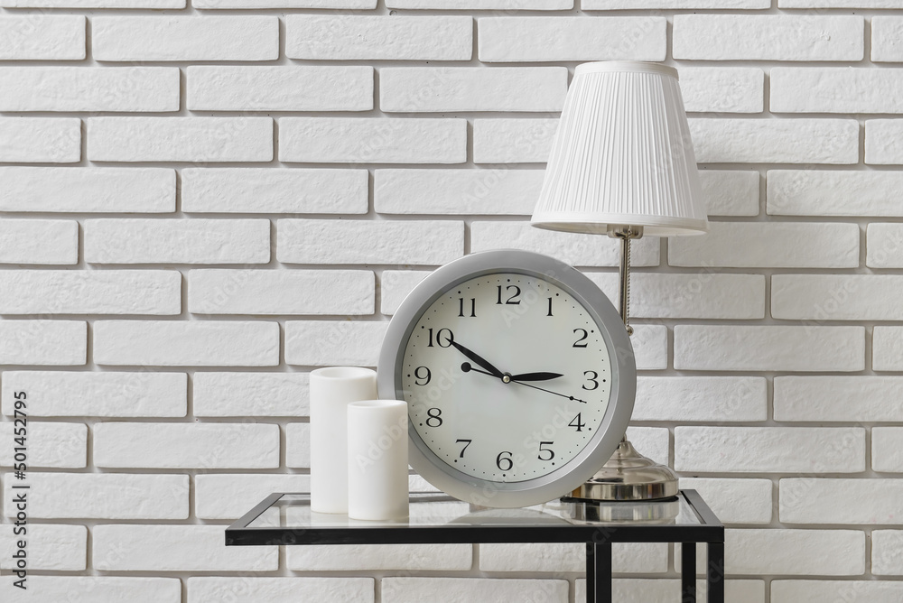 Table with modern clock, lamp and candles near light brick wall in room