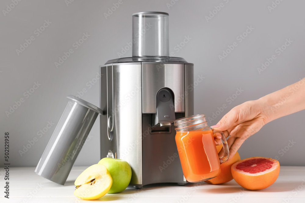 Female hand holding mason jar of fresh juice with fruits and new modern juicer on table