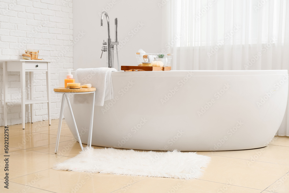 Modern bathtub with tray and different supplies on table in bathroom interior