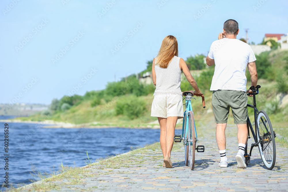 Mature couple with bicycles walking along river bank on summer day