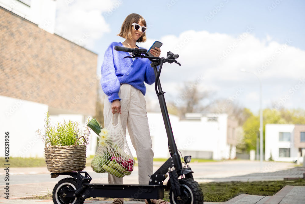 Happy stylish woman using phone while stannding with fresh vegetables and greens near electrical sco