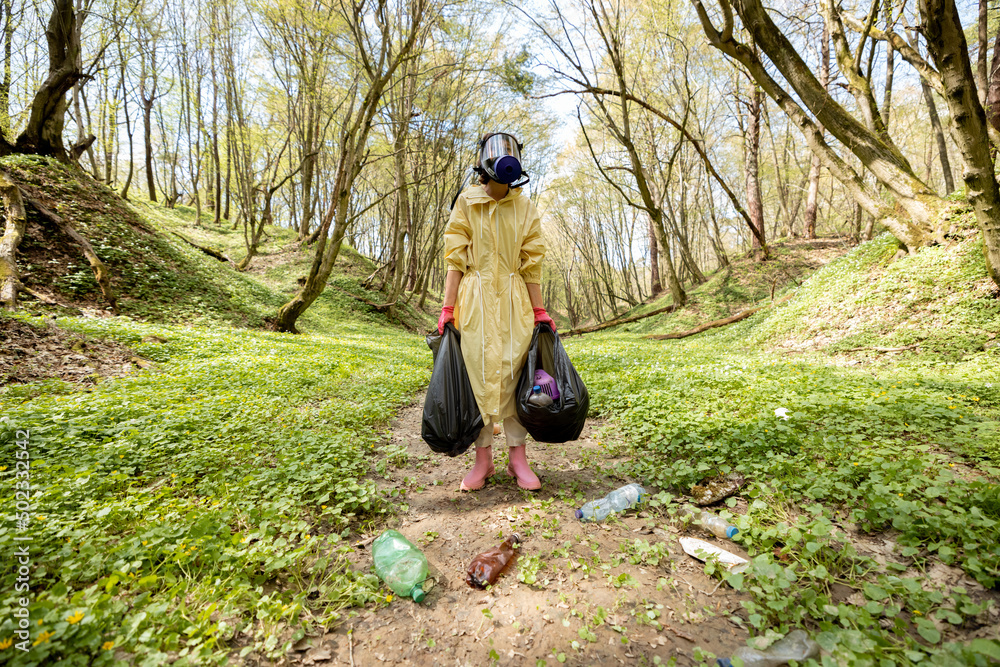 Woman in gas mask and protective clothes stands with garbage bags collecting scattered plastic in th