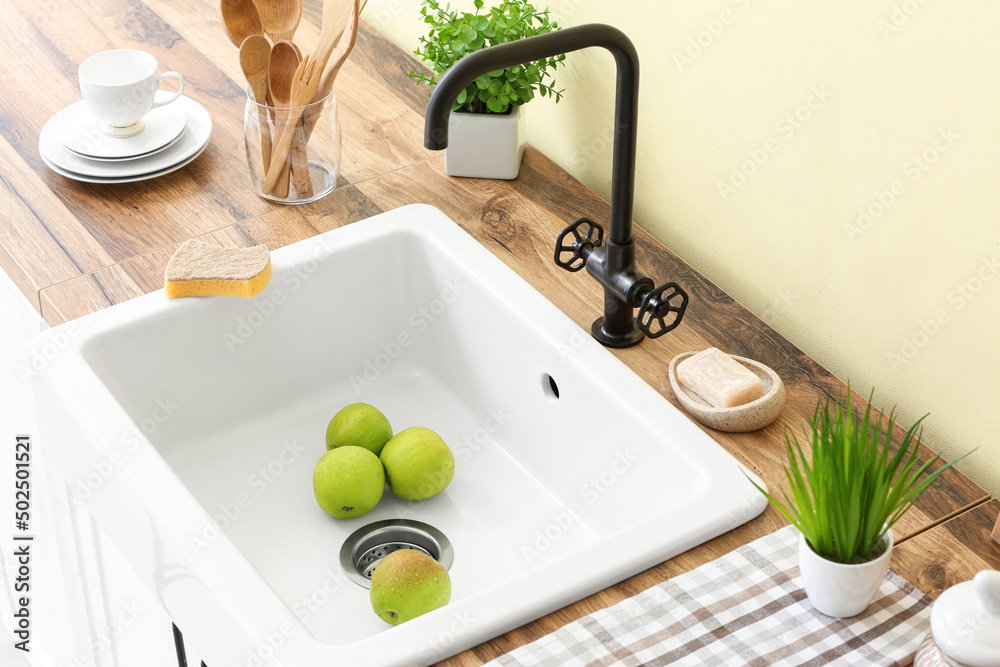 Fresh green apples in sink and different dishware on wooden counter near color wall