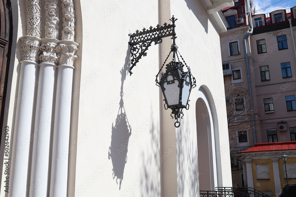 The shadow of a large vintage street lamp falls on the wall on a sunny day. St. Annas Church, Russi