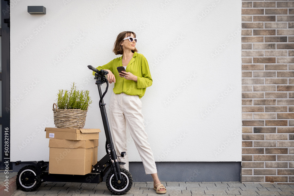 Woman standing with phone near electric scooter with some parcels and flowerpot on a porch of her ho