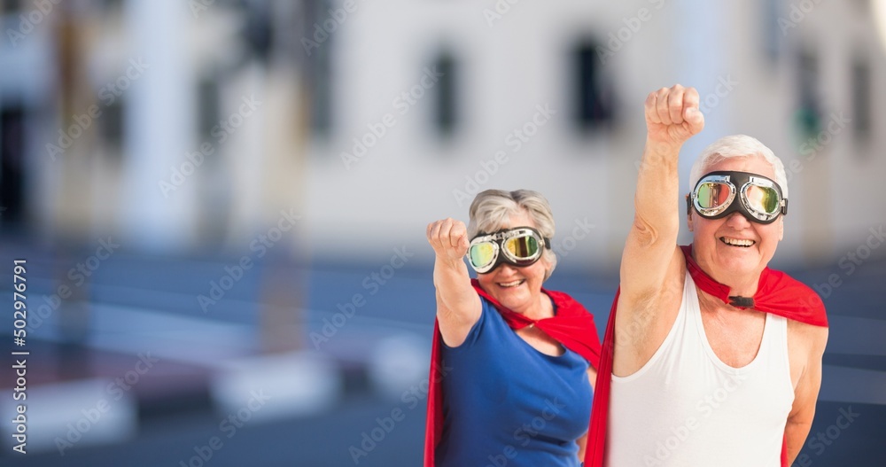 Happy caucasian senior couple wearing red capes and masks with hand raised standing in city